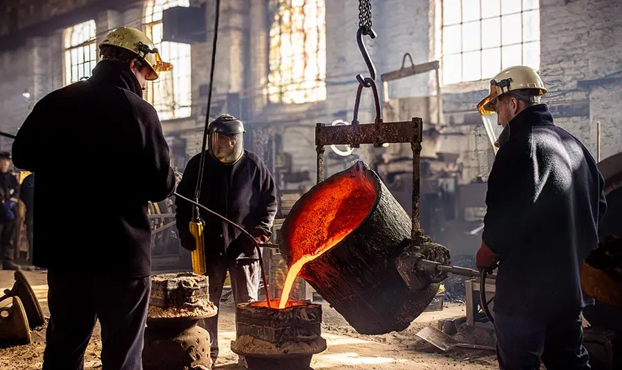 loughborough_bell_foundry_bell_casting_ckatelyn_collins_web_size.jpg Three people stand inside a factory casting a large bell. Molten metal pours from a container held in the air by a hoist